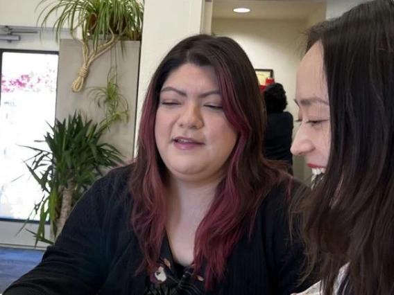 Two women sitting at a table looking at a computer. One woman is smiling and the other is speaking.