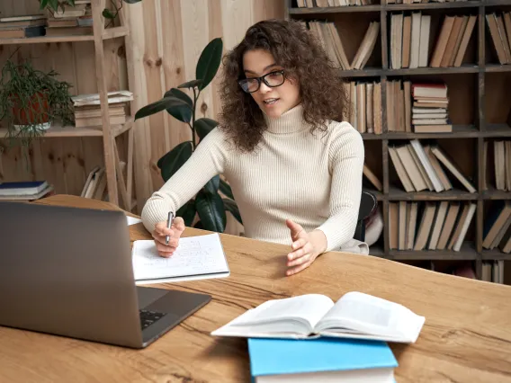 A woman in front of a bookshelf speaking to a laptop